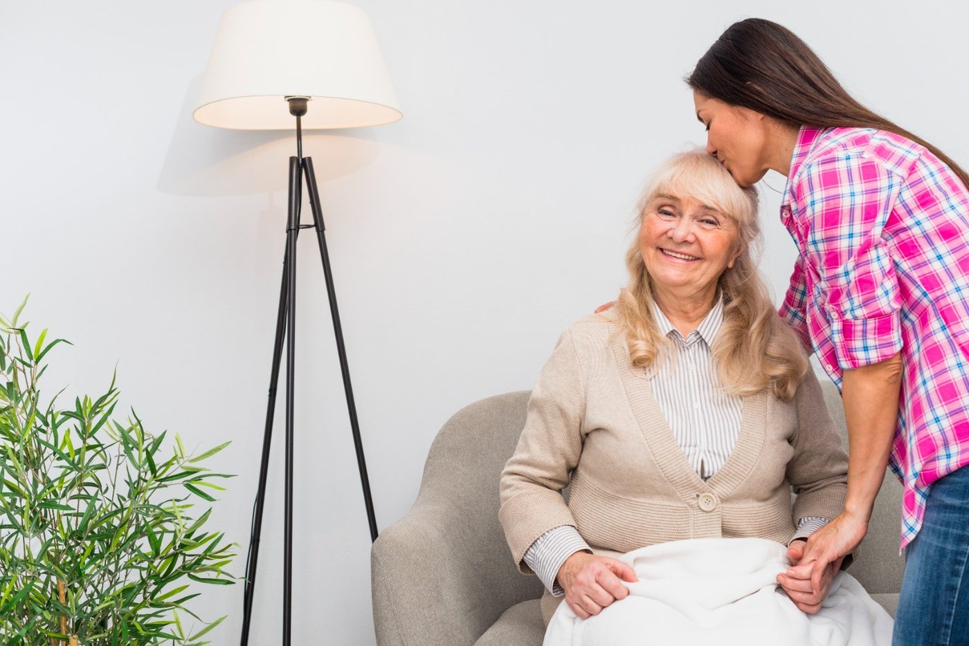 A young woman kissing a smiling older woman on the head as she sits in her armchair