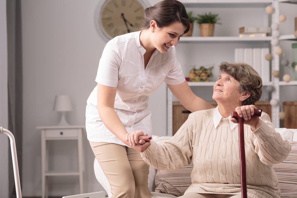 A live-in carer helping an elderly woman with a walking stick to stand