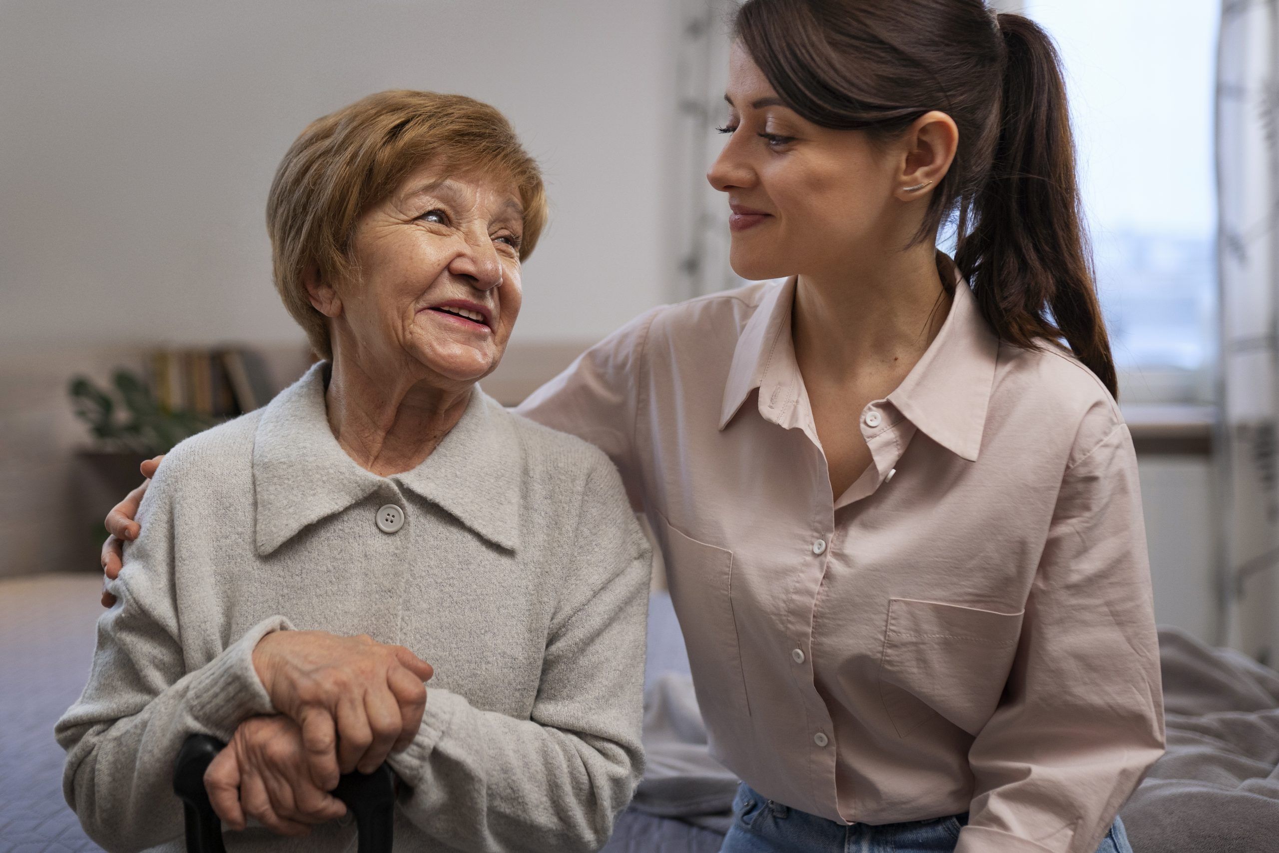 A live-in carer with her arm around a smiling older woman holding her walking stick at home